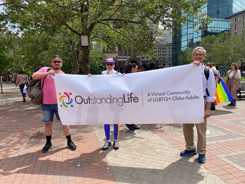 Three people holding an Outstanding Life banner and marching with/for LGBTQ+ elders at Boston Pride